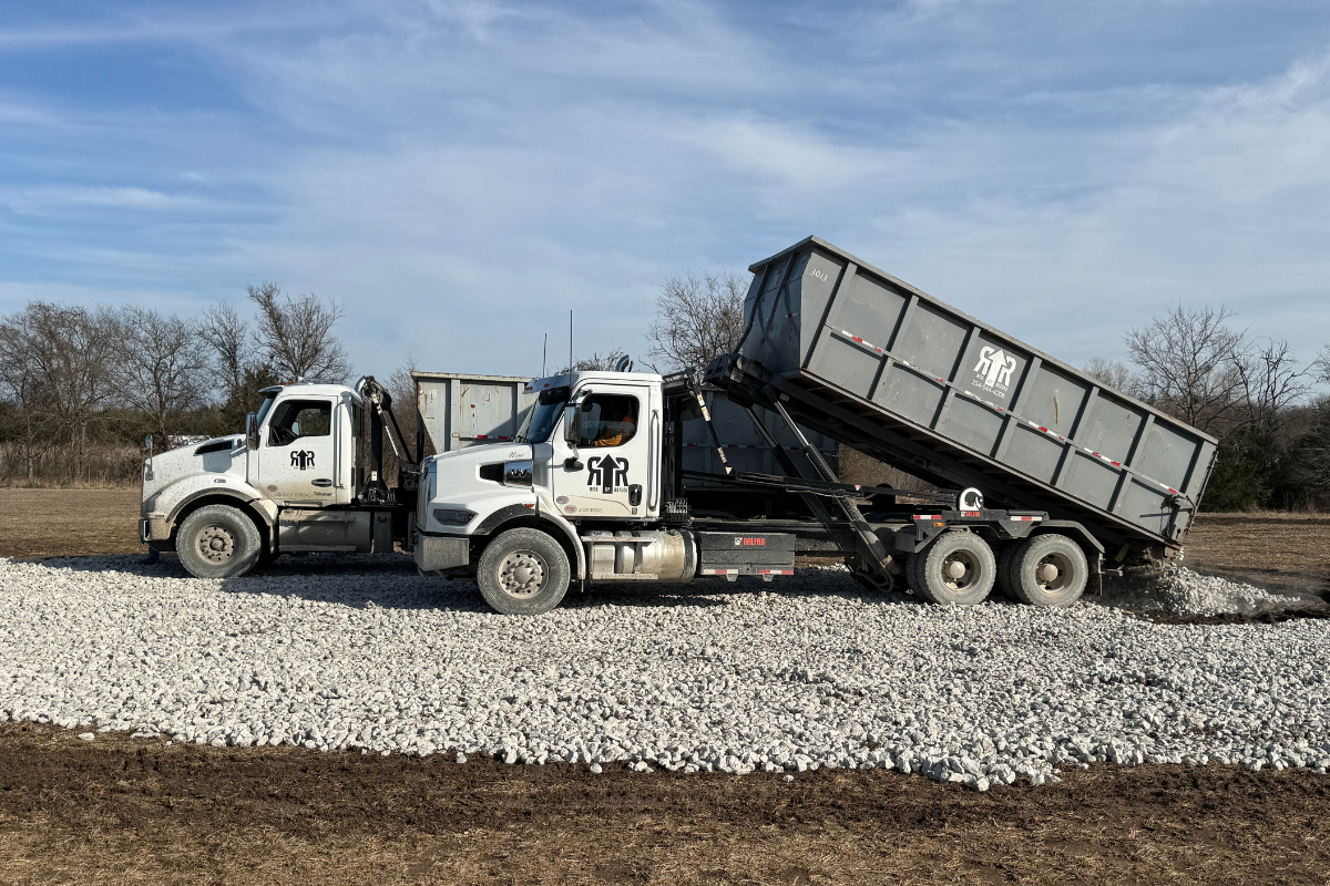 Two trucks with roll-off dumpsters dumping rocks in an empty field