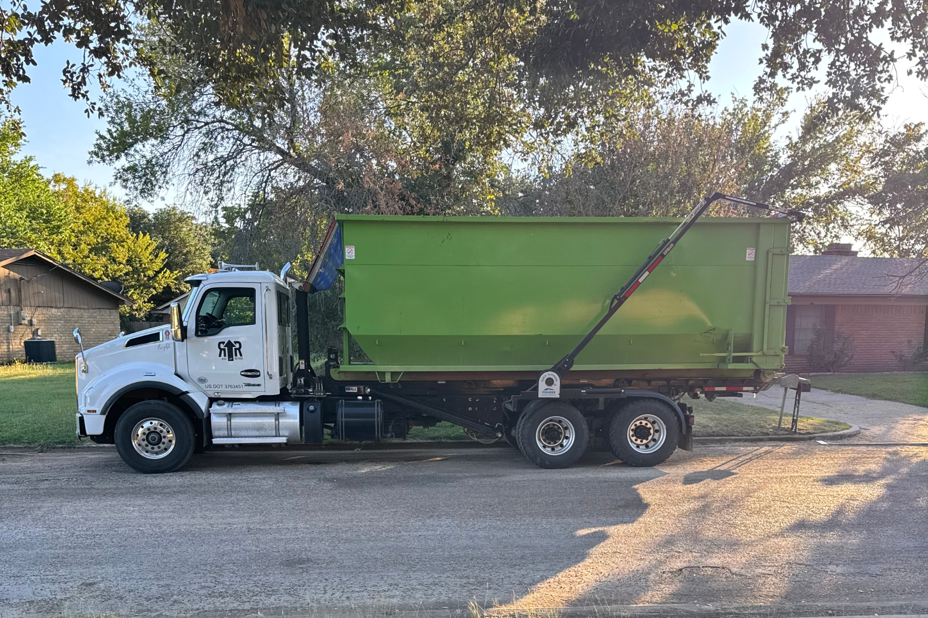 Dump truck carrying green dumpster in front of residential home